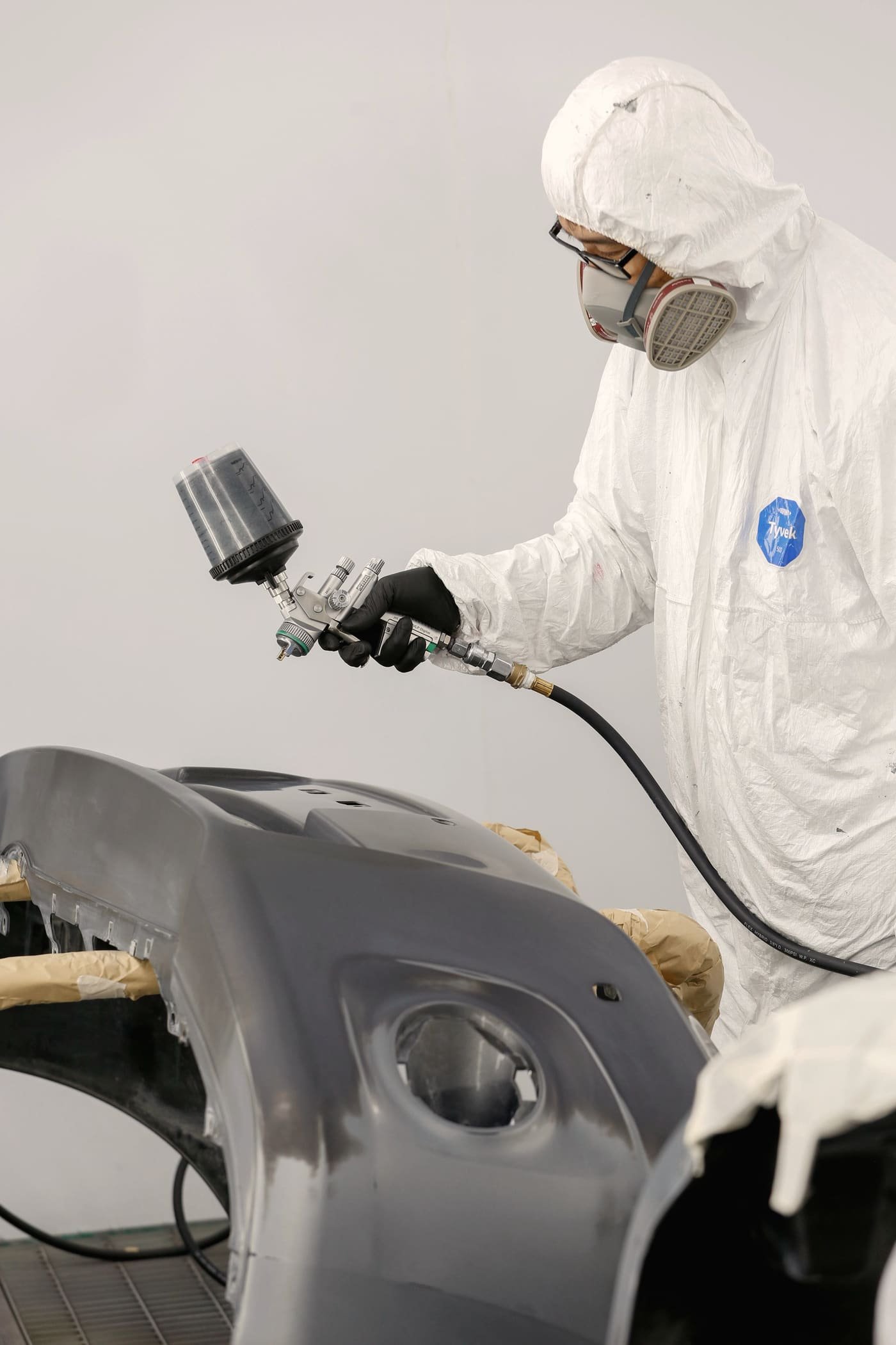 Painter in a full suit and respirator spraying a primered bumper cover on a stand